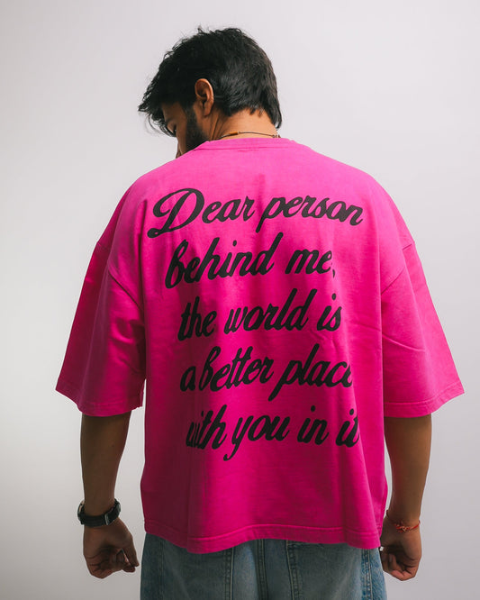 A person viewed from behind wearing a bright pink oversized T-shirt with black cursive text on the back reading, 'Dear person behind me, the world is a better place with you in it,' against a plain light gray background. Positive, feel good t shirt